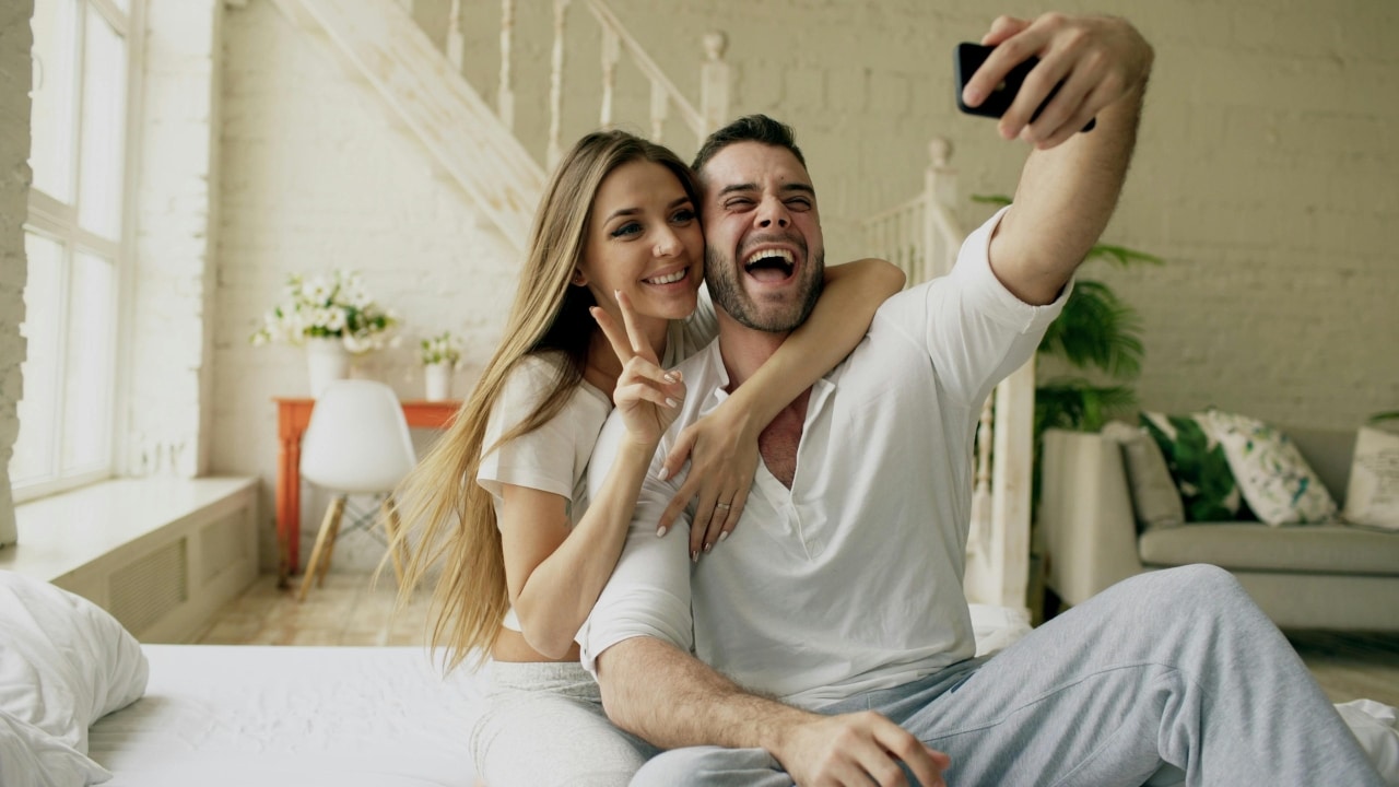 A woman hugs a man from behind as he holds a phone to take a smiling selfie.