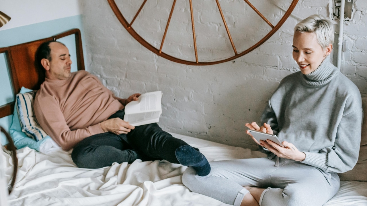 A man reads a book and a woman uses a tablet while sitting on bed.