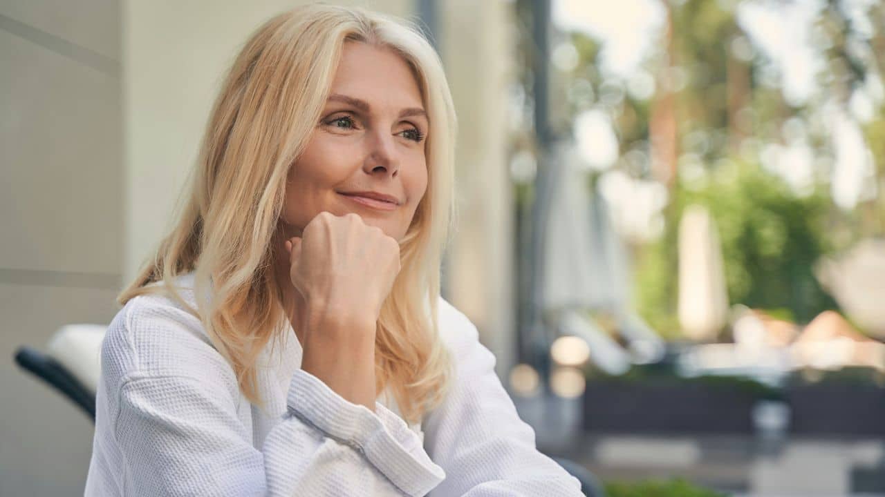 A blonde woman in a white robe rests her chin on her hand and smiles.