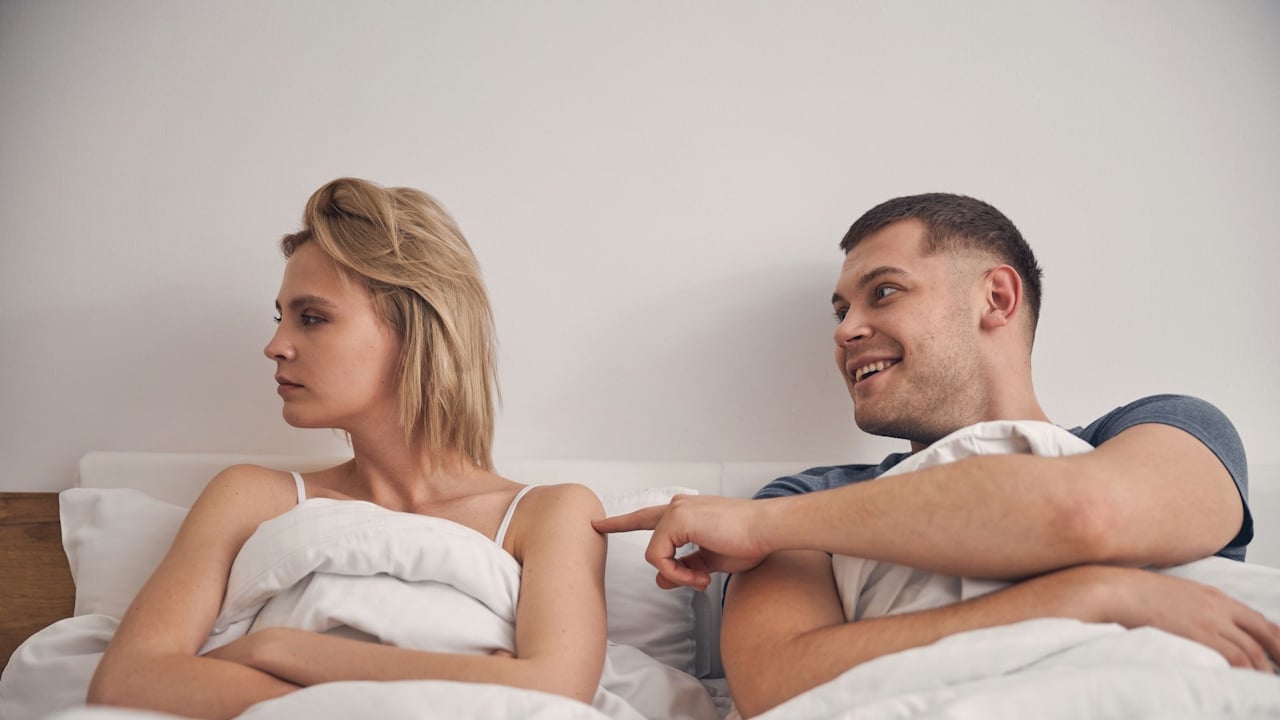 A blonde woman with her arms crossed is looking away from a man who is smiling and trying to get her attention by poking her back in bed.