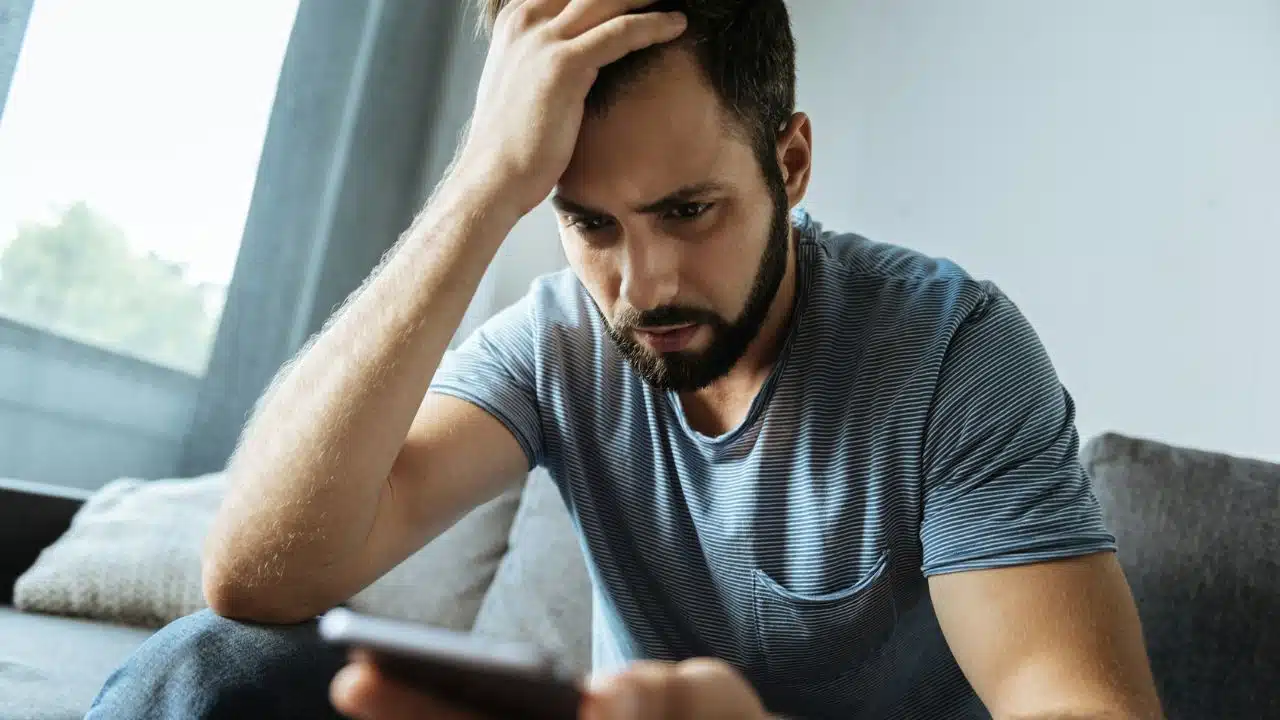 A bearded man sits on a couch, holding his head while looking at a smartphone.