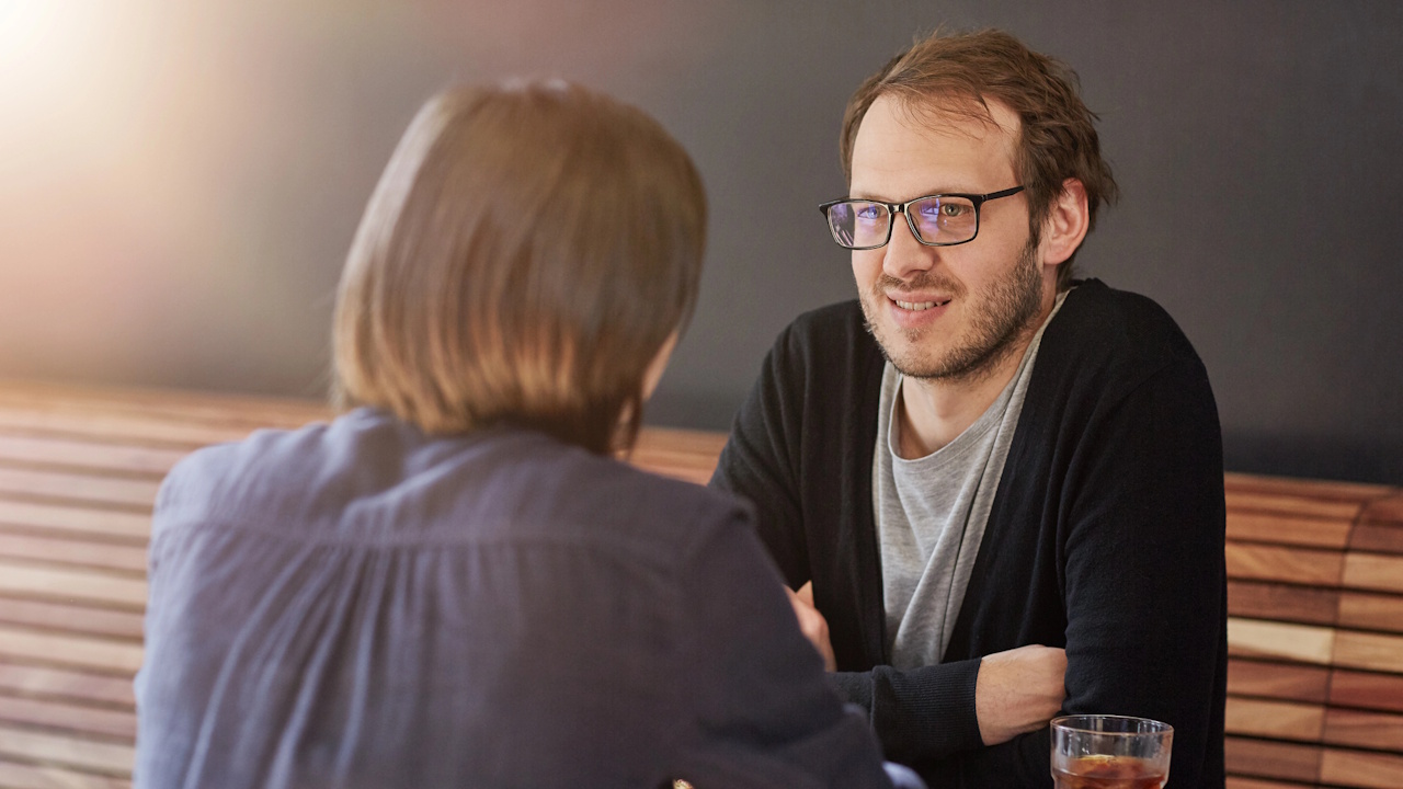 A man in glasses smiling across a table at another person.