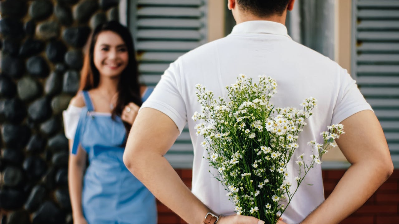 A man holding a bouquet of white flowers behind his back, surprising a smiling woman.