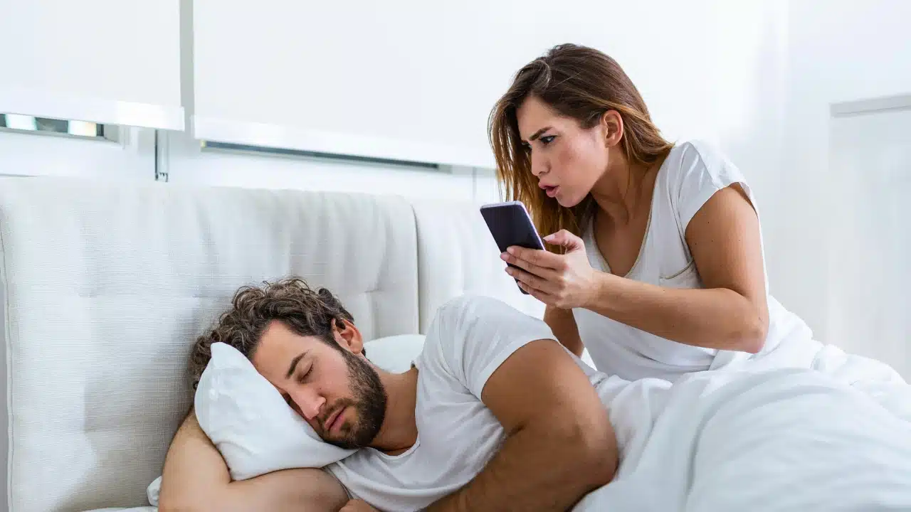 A woman looks at a smartphone with an angry expression while a man sleeps nearby.