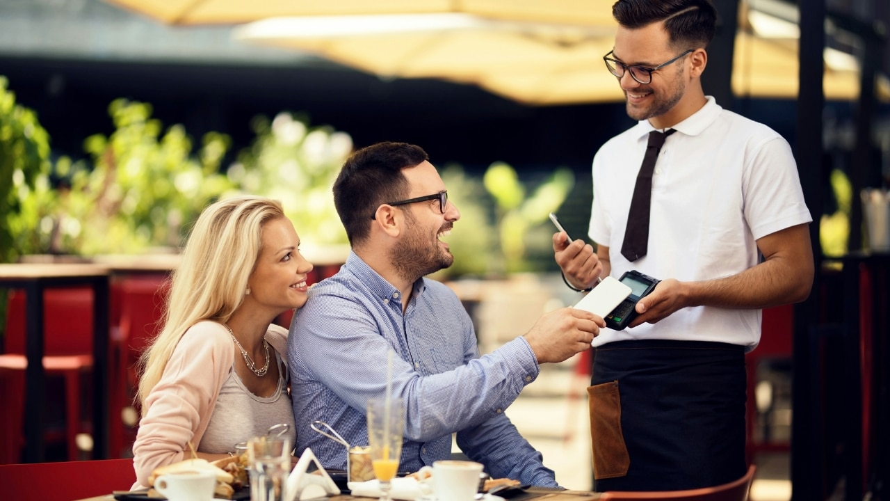 A man at an outdoor table pays a server using a mobile phone and terminal.
