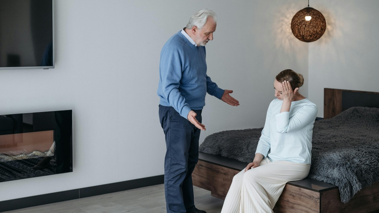 An older man stands and gestures while a woman sits on a bed looking down.