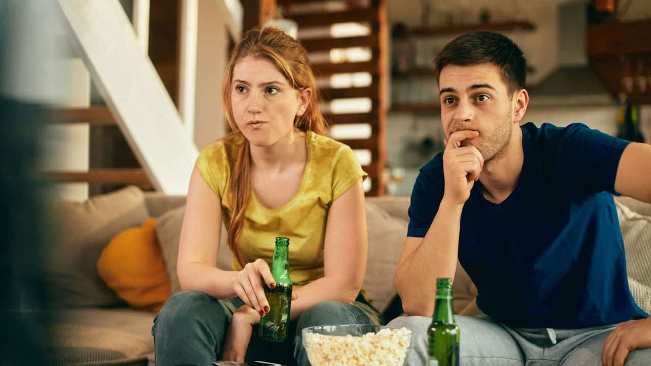 A man and woman sitting on a couch, holding beer bottles and watching television.