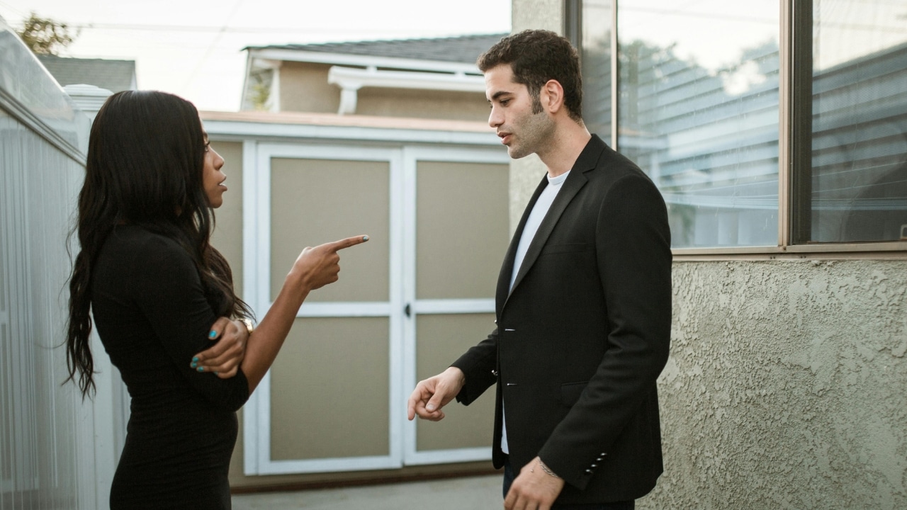 A woman points her finger at a man while they stand outside near a building.