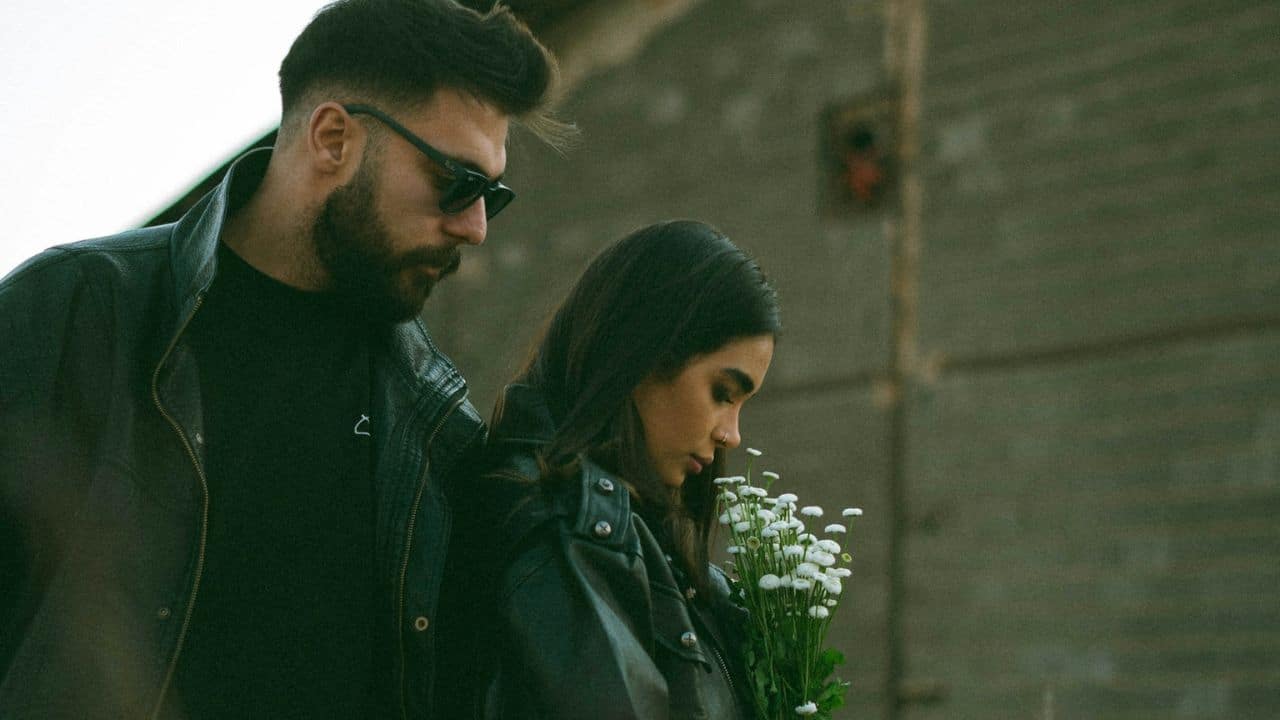 A bearded man in sunglasses stands behind a woman holding a bunch of white flowers.