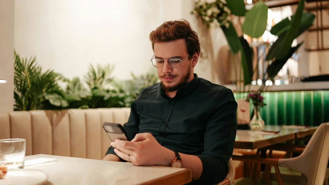 A man with glasses and a beard sits at a restaurant table using a smartphone.