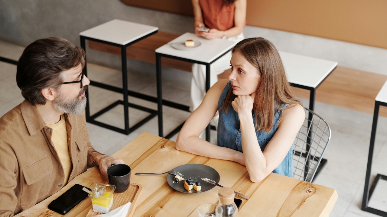 A man and woman sit at a wooden table with food and drinks, looking at each other.