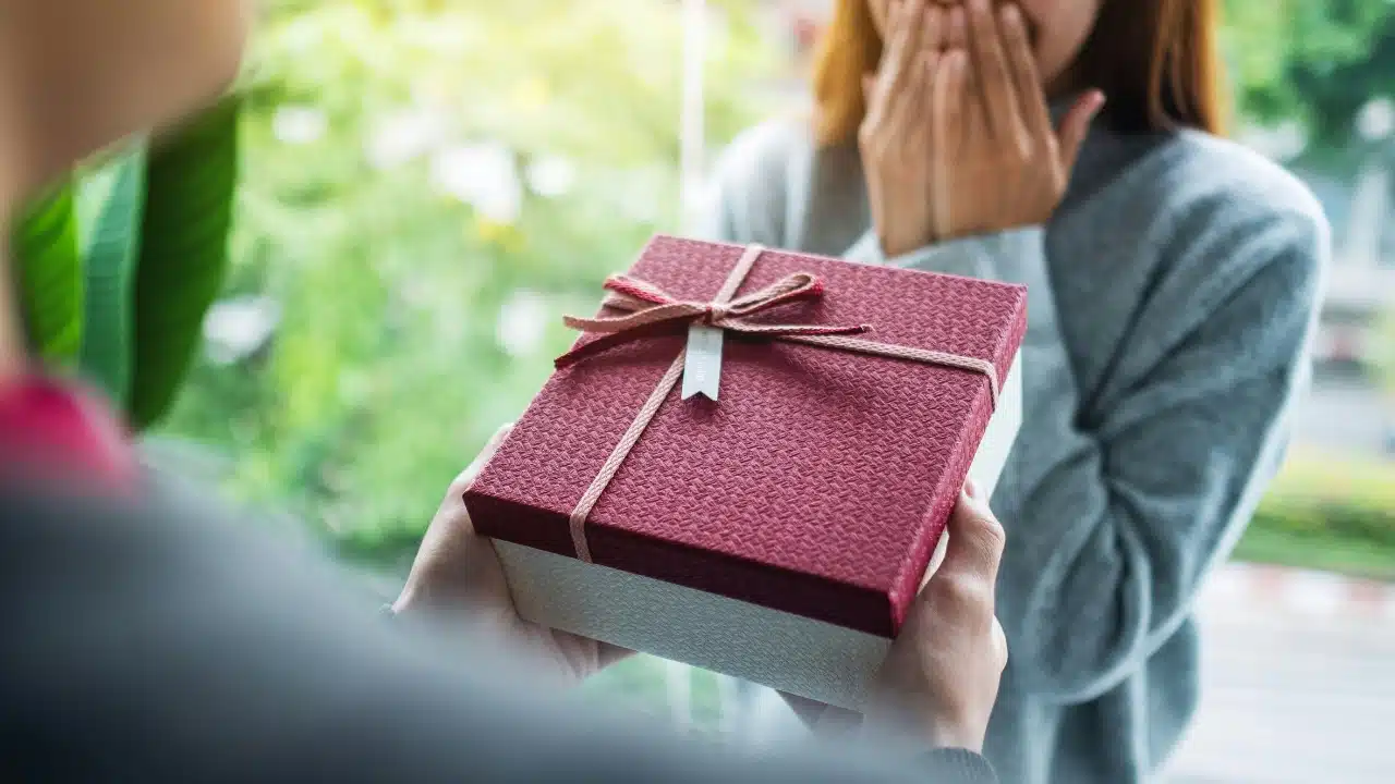A person holds out a red and white gift box toward a woman covering her mouth.