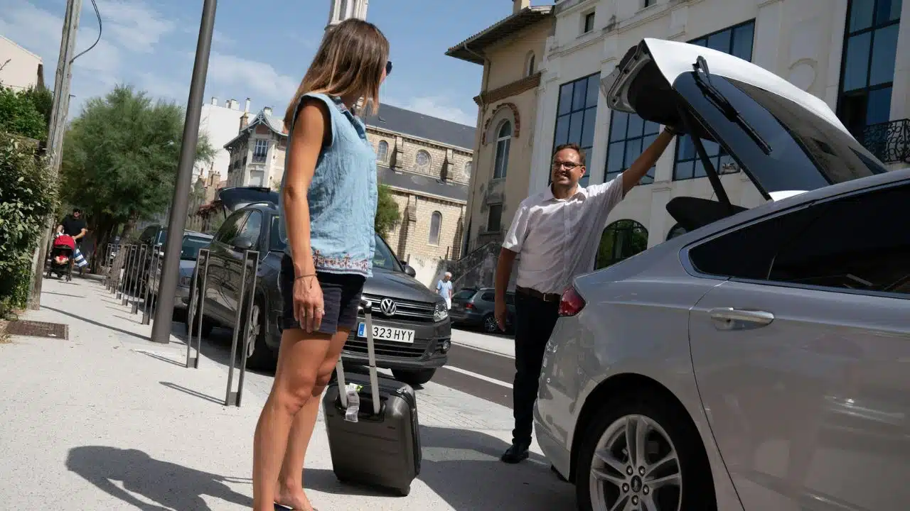 A woman stands with a suitcase while a man opens the trunk of a car.