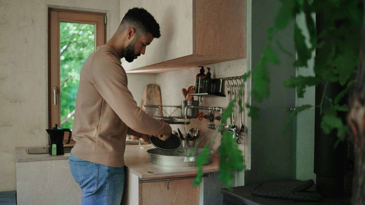 A man with a beard stands in a kitchen washing a pan in a metal sink.