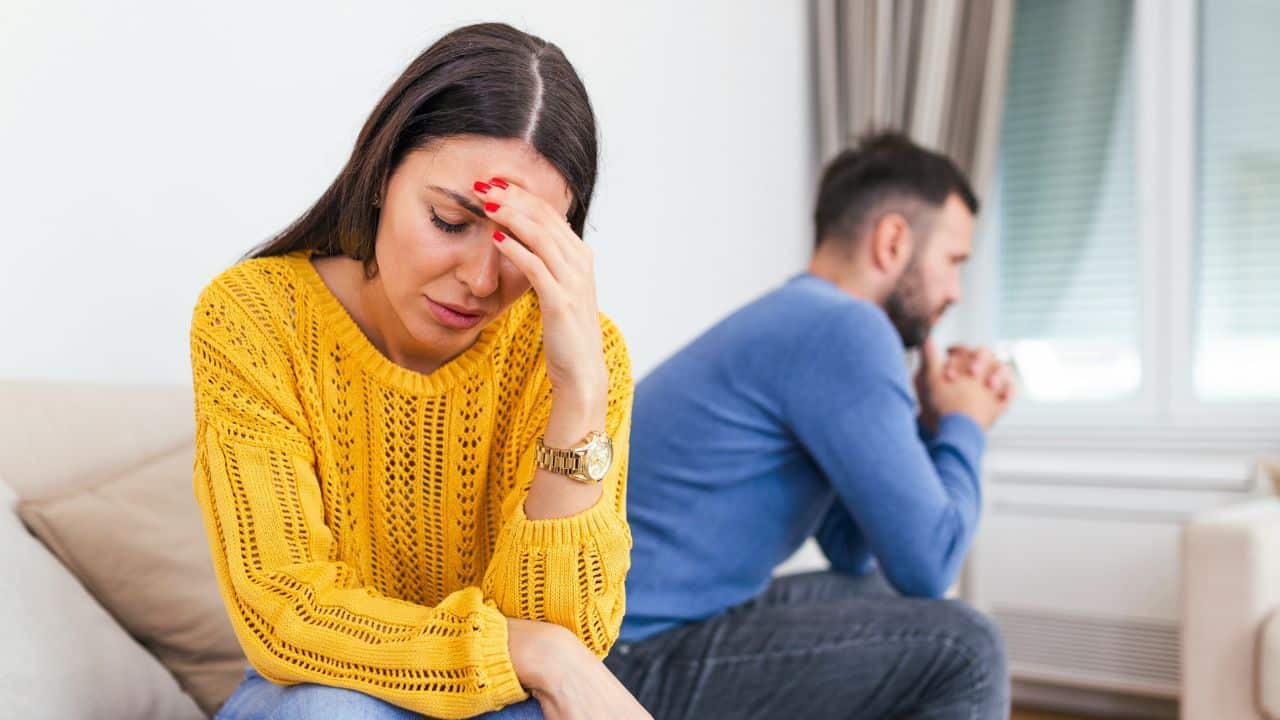 A woman in a yellow sweater holds her forehead while a man sits behind her.