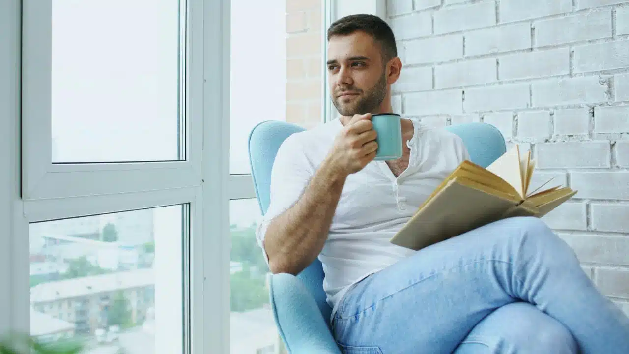 A man sits in a blue chair by a window, holding a mug and a book.