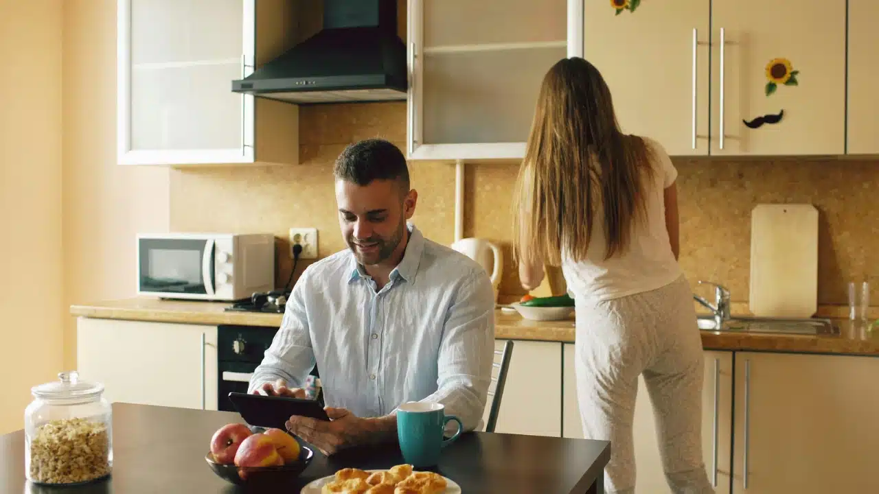 A man uses a tablet at a table while a woman works in the kitchen background.