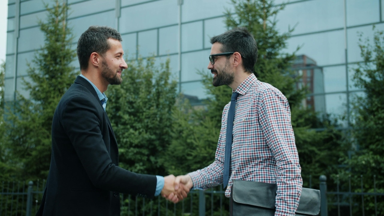 Two men in professional attire shake hands outdoors in front of a glass building and trees.