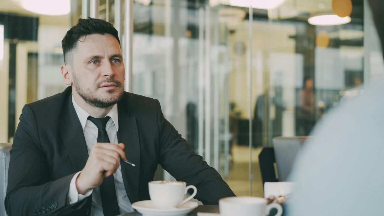 A man in a dark suit and tie sits at a table with coffee cups.