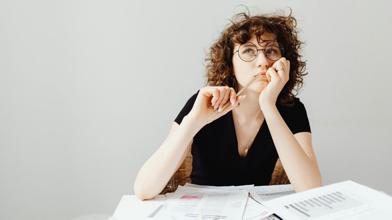 A woman with curly hair and glasses holds a pencil to her chin while looking at paperwork.