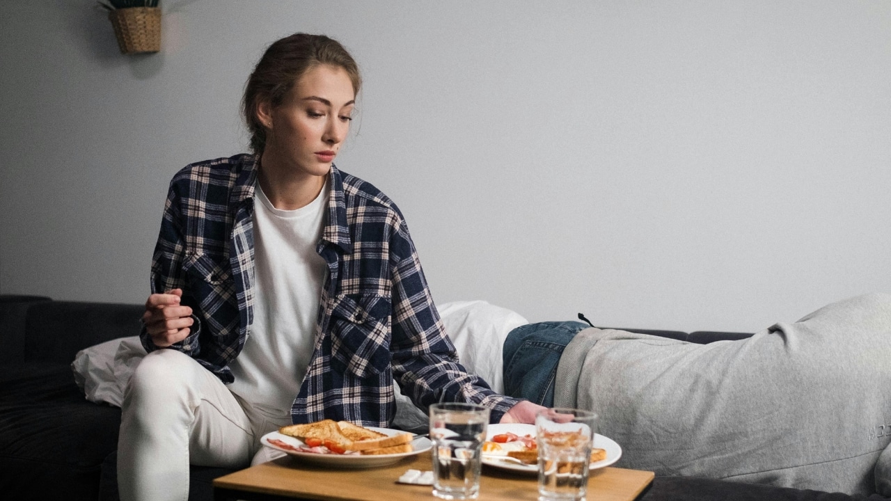 A woman in a plaid shirt sits by a table with breakfast near a person.
