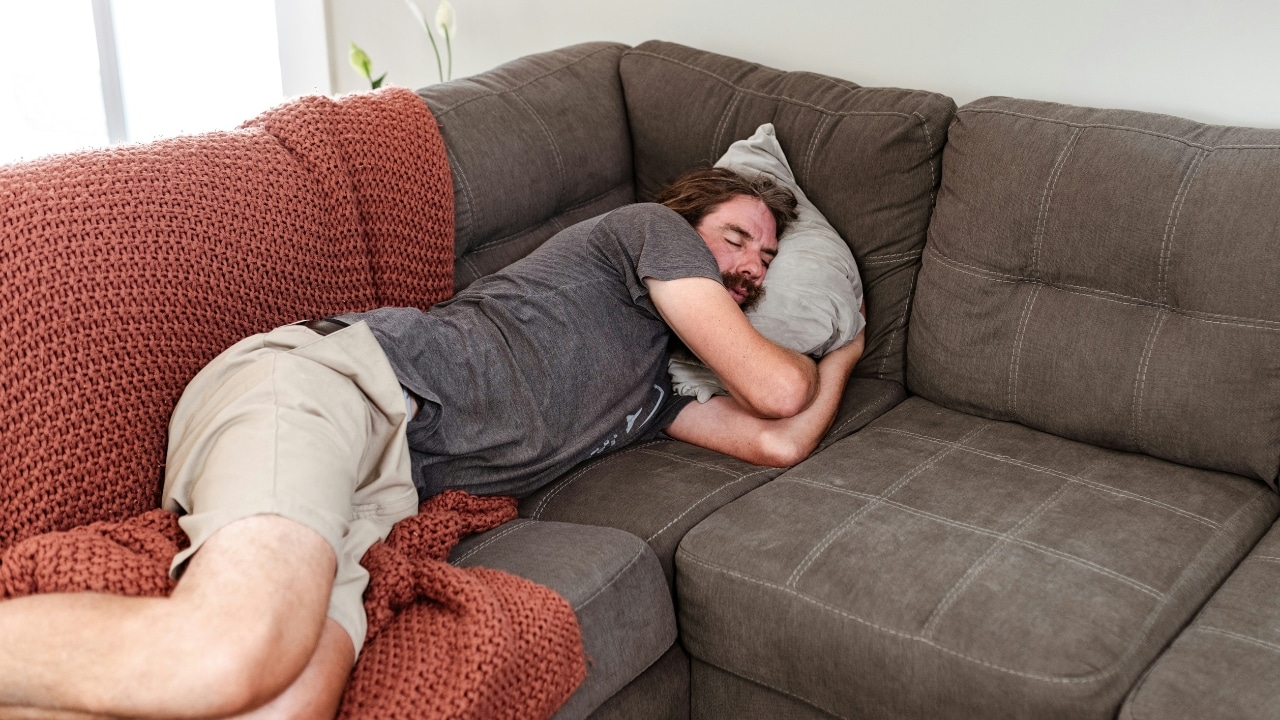 A man sleeps on his side on a brown couch tucked under a red blanket.