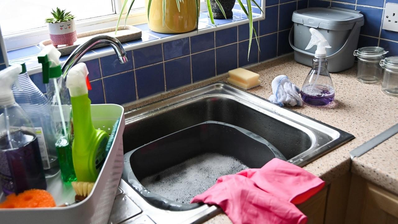 A kitchen sink filled with soapy water sits next to cleaning supplies and pink gloves.