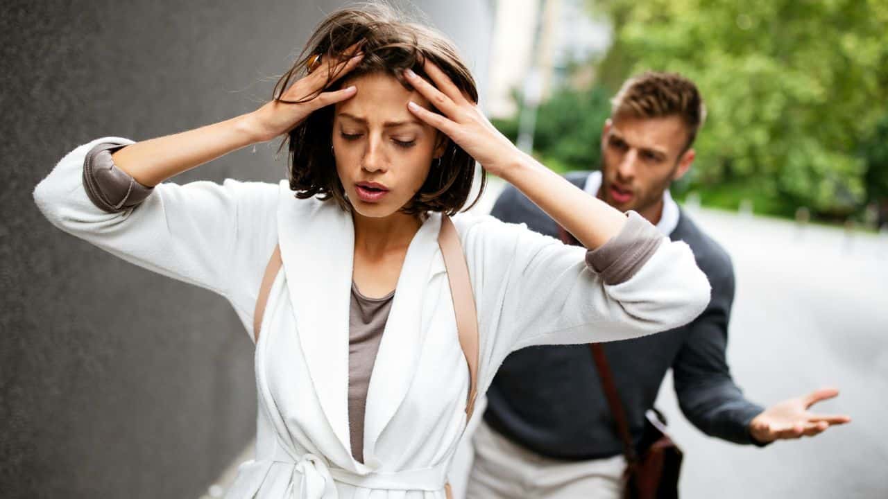 A woman holds her head with both hands while a man gestures behind her outdoors.