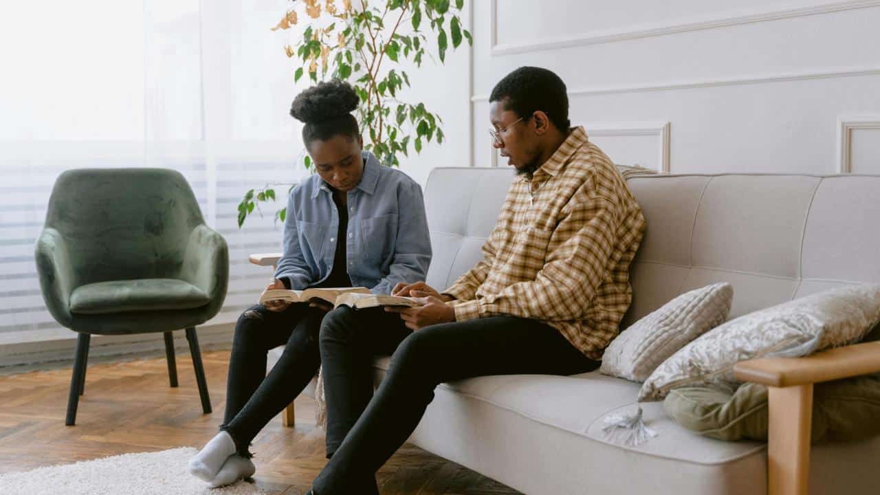 A couple sitting on a couch reading books together in a bright living room.