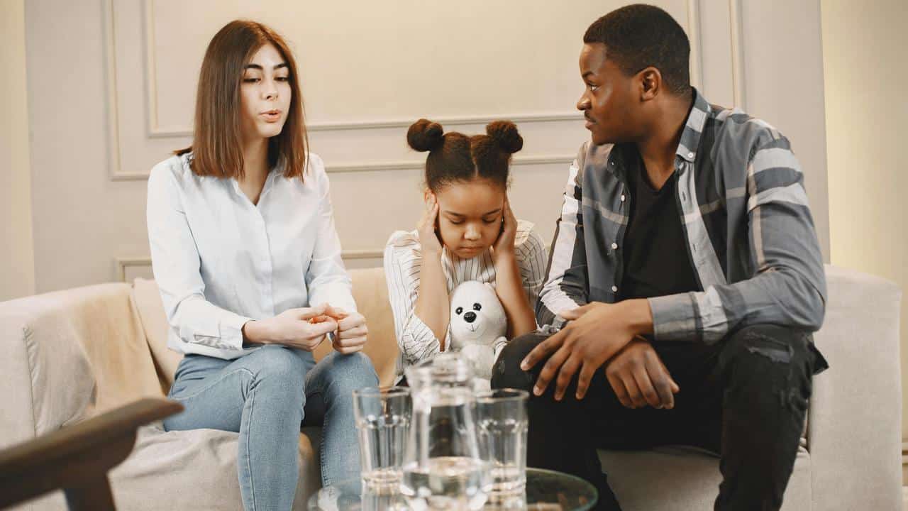 A couple with a young girl holding a stuffed toy during a serious conversation.