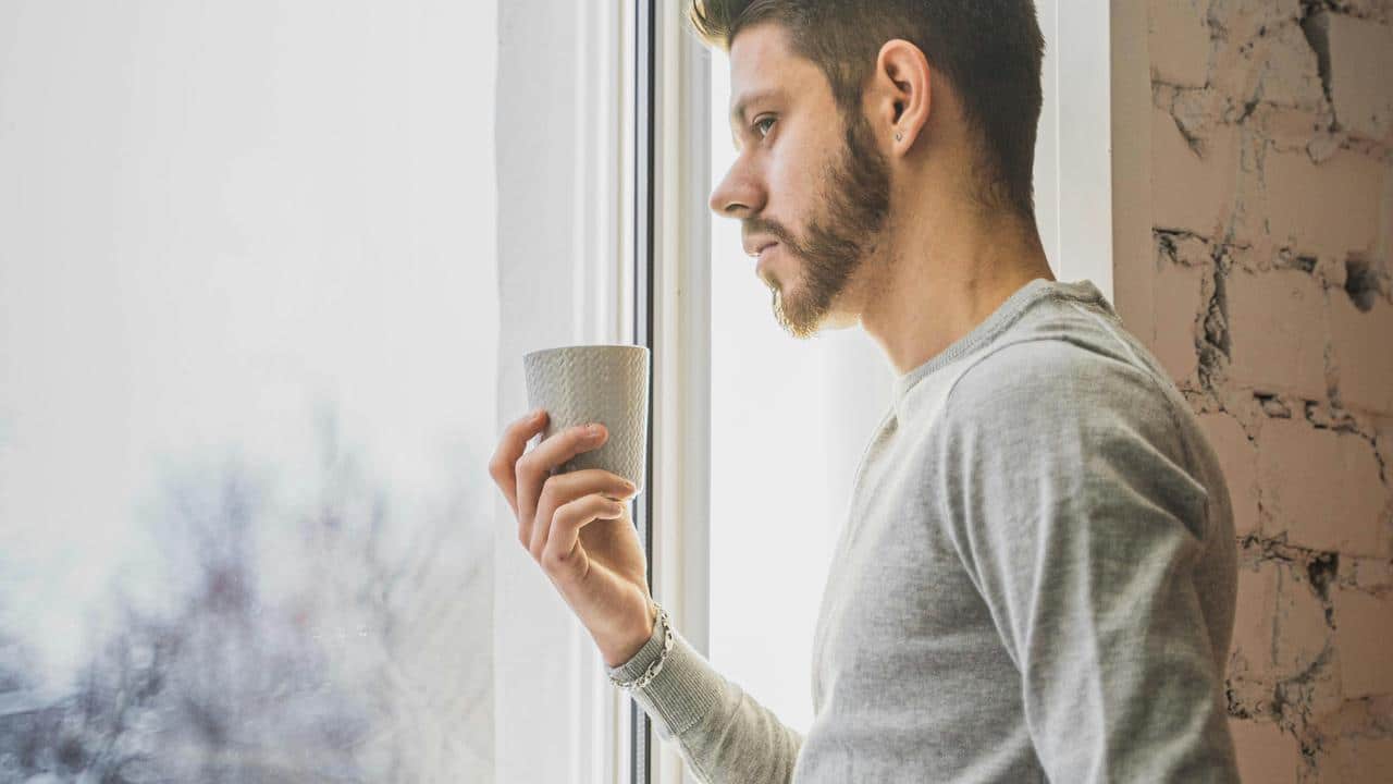 A man standing by a window holding a cup, looking outside thoughtfully.