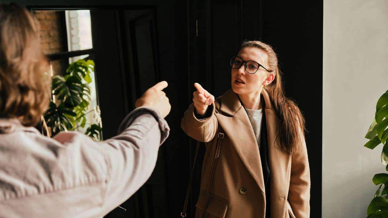 A woman in a tan coat and glasses points angrily at a man.