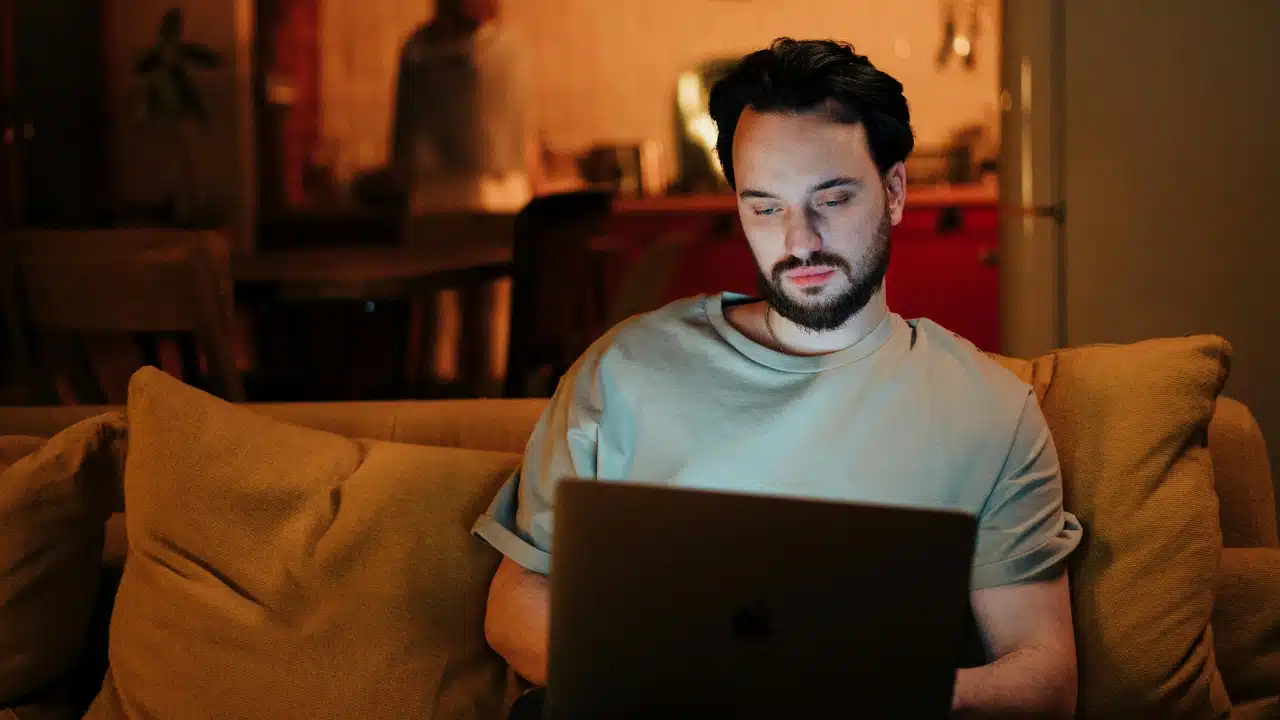 A bearded man sits on a couch at night looking at a glowing laptop screen.