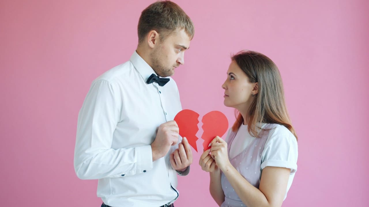 A couple holding broken-heart halves on a pink background.