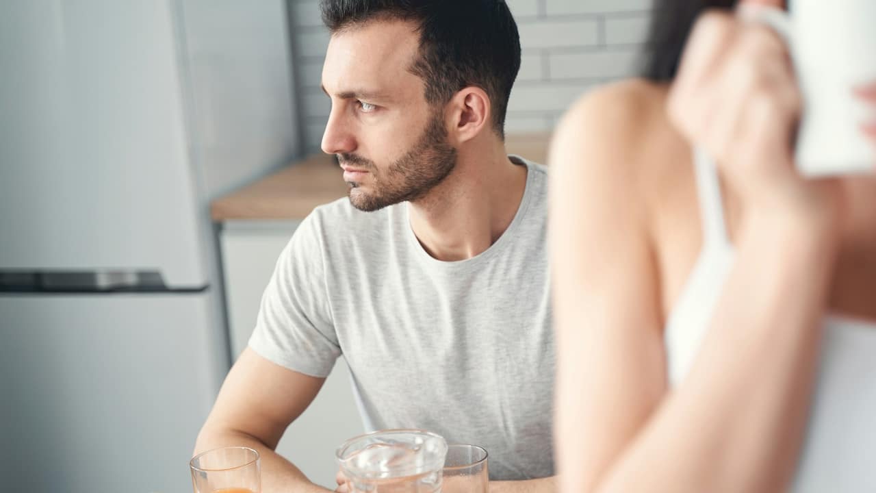 A lightly bearded and angry man looking to the side while a woman sips coffee.