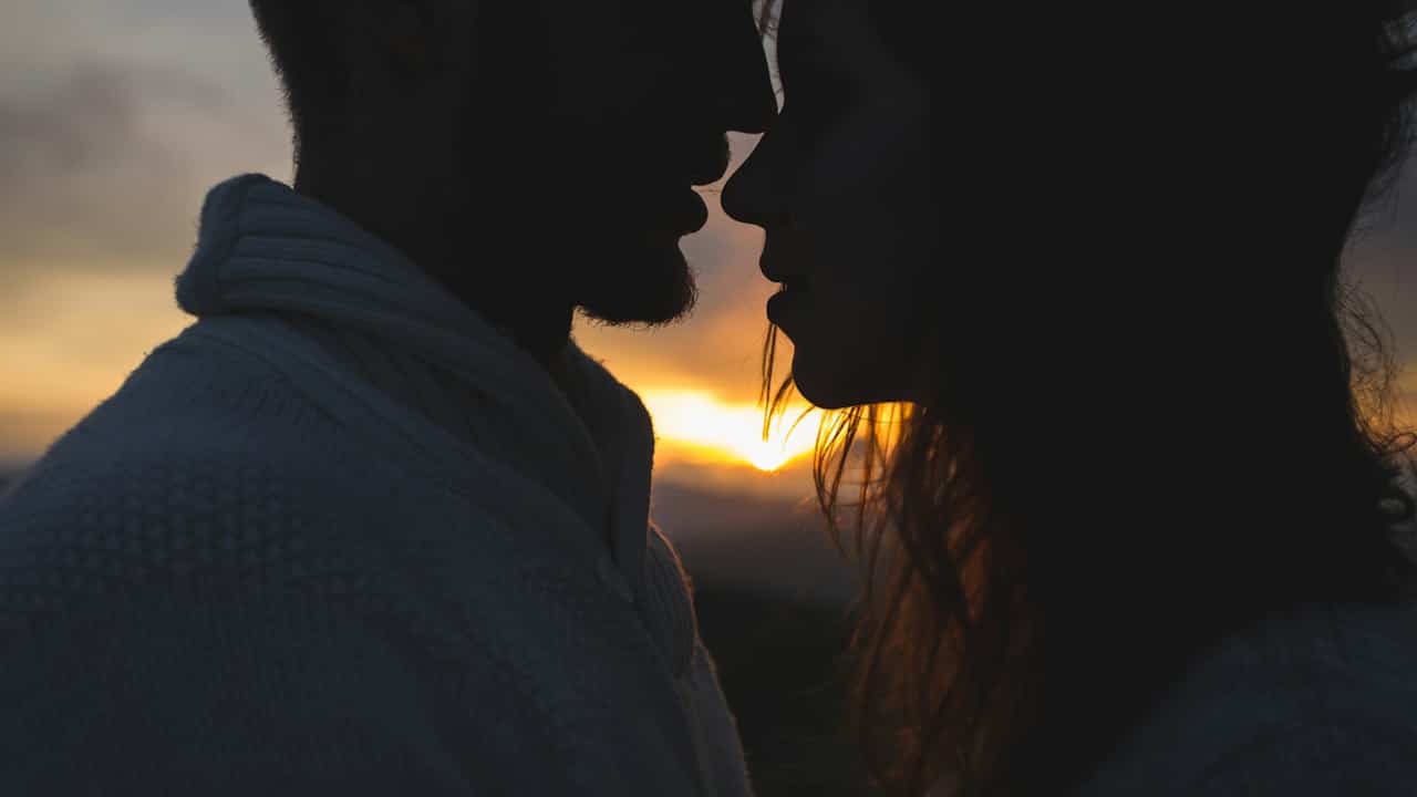 A romantic silhouette of a couple standing nose-to-nose, about to kiss, with the sunset glowing brightly between them.