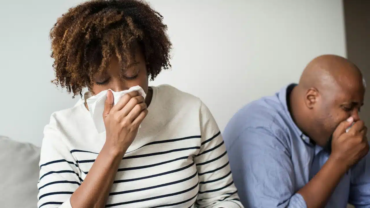 A woman and a man sit side by side, woman blowing her nose with tissue.
