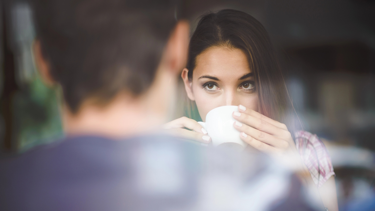 A woman looking up while drinking coffee across from a blurred man.