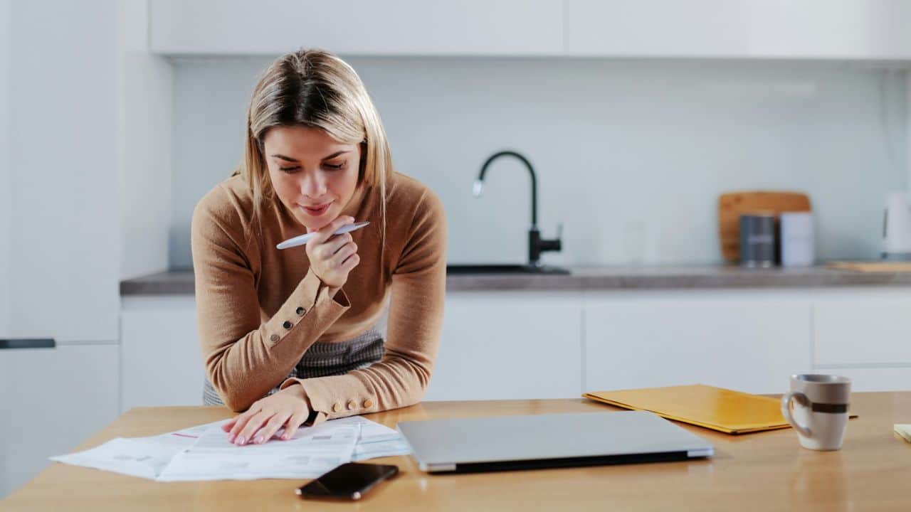 A woman leans over a wooden table looking at papers next to a closed laptop.