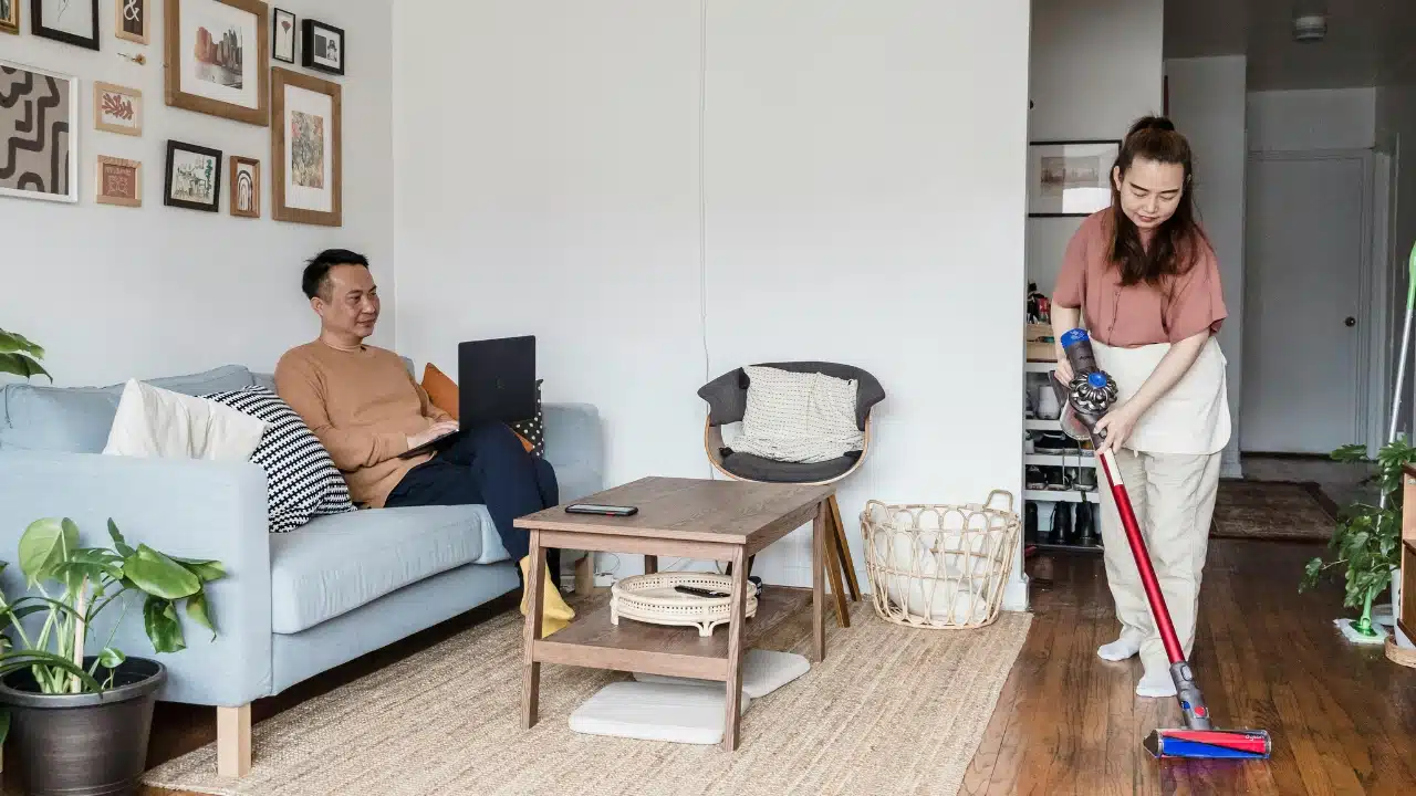 A woman uses a vacuum cleaner while a man sits on a sofa with a laptop.