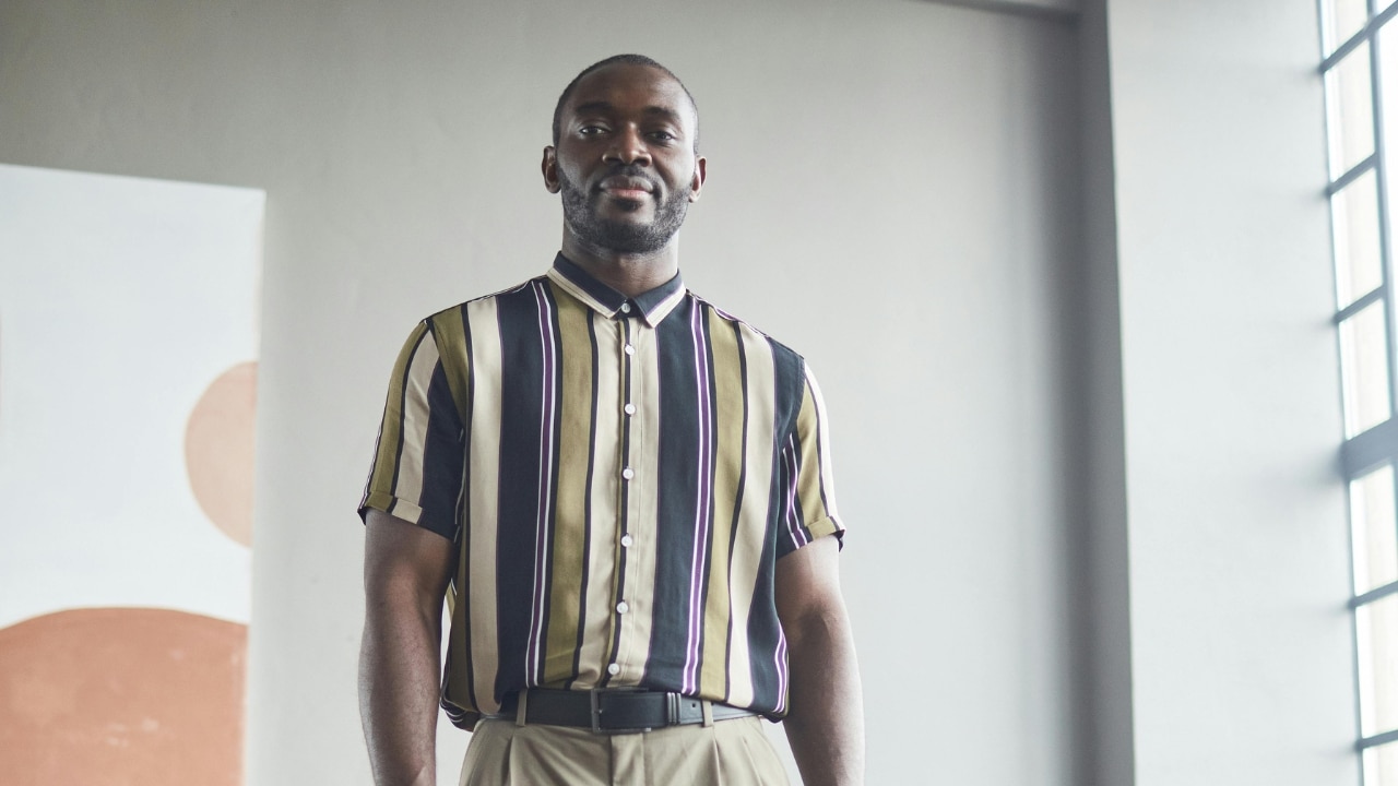 A man wearing a striped short-slee shirt and khaki trousers stands against a gray wall.