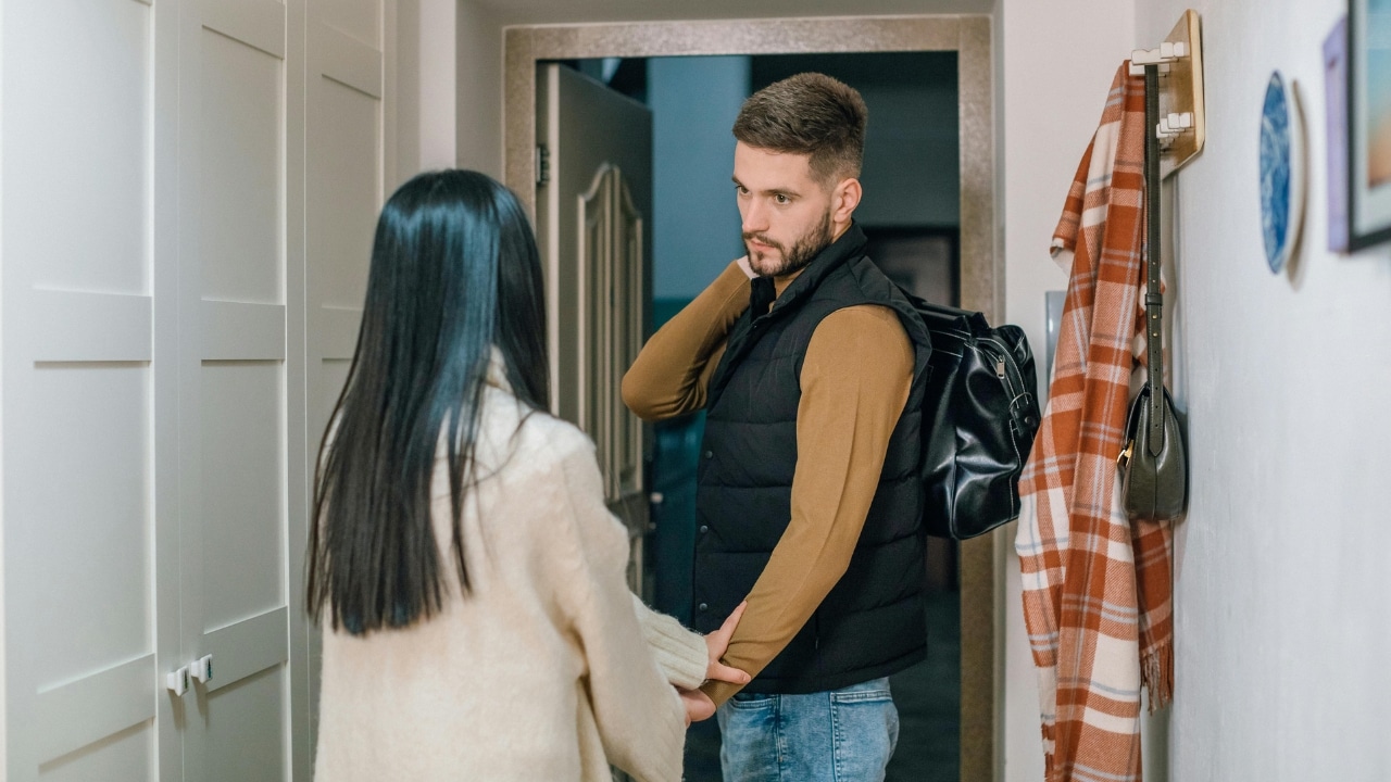 A man carrying a black bag looks at a woman standing in a hallway.