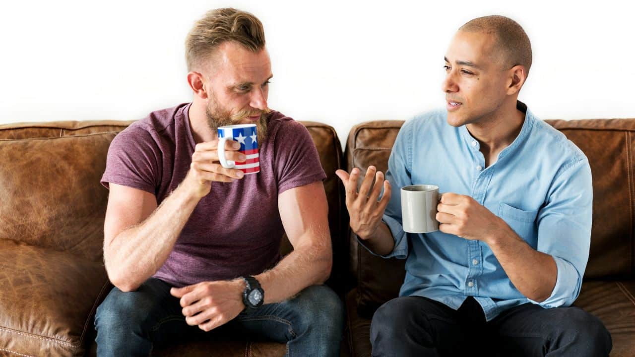 Two men sit on a leather sofa holding mugs while engaged in a serious conversation.