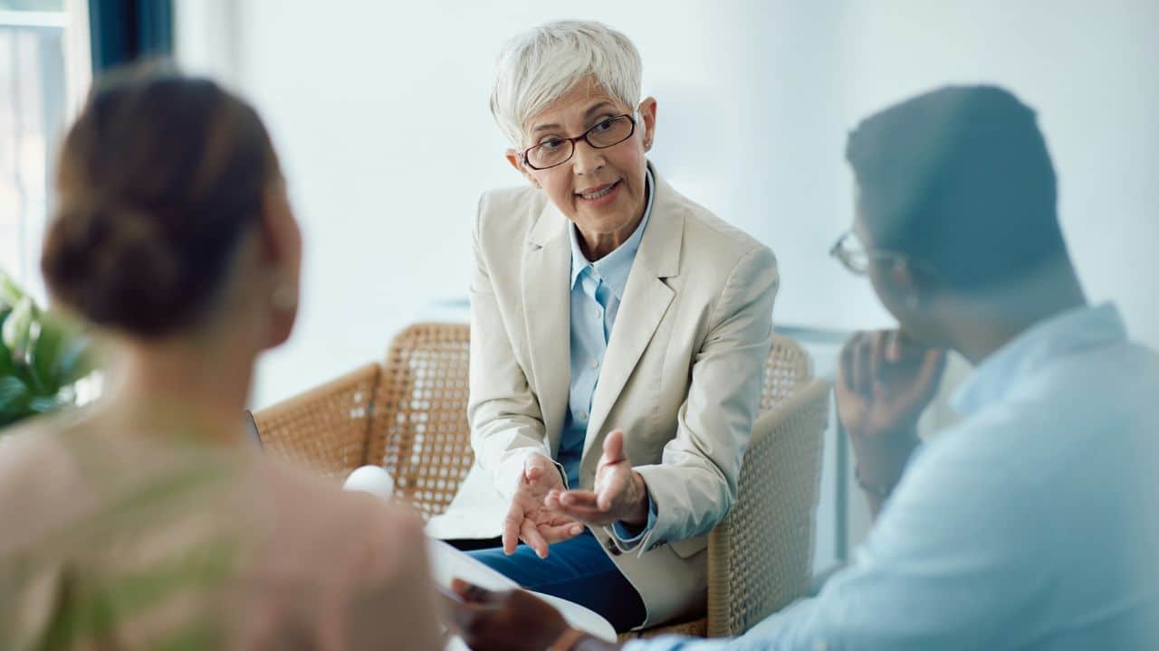 A woman with short grey hair and glasses speaks to two people sitting before her.