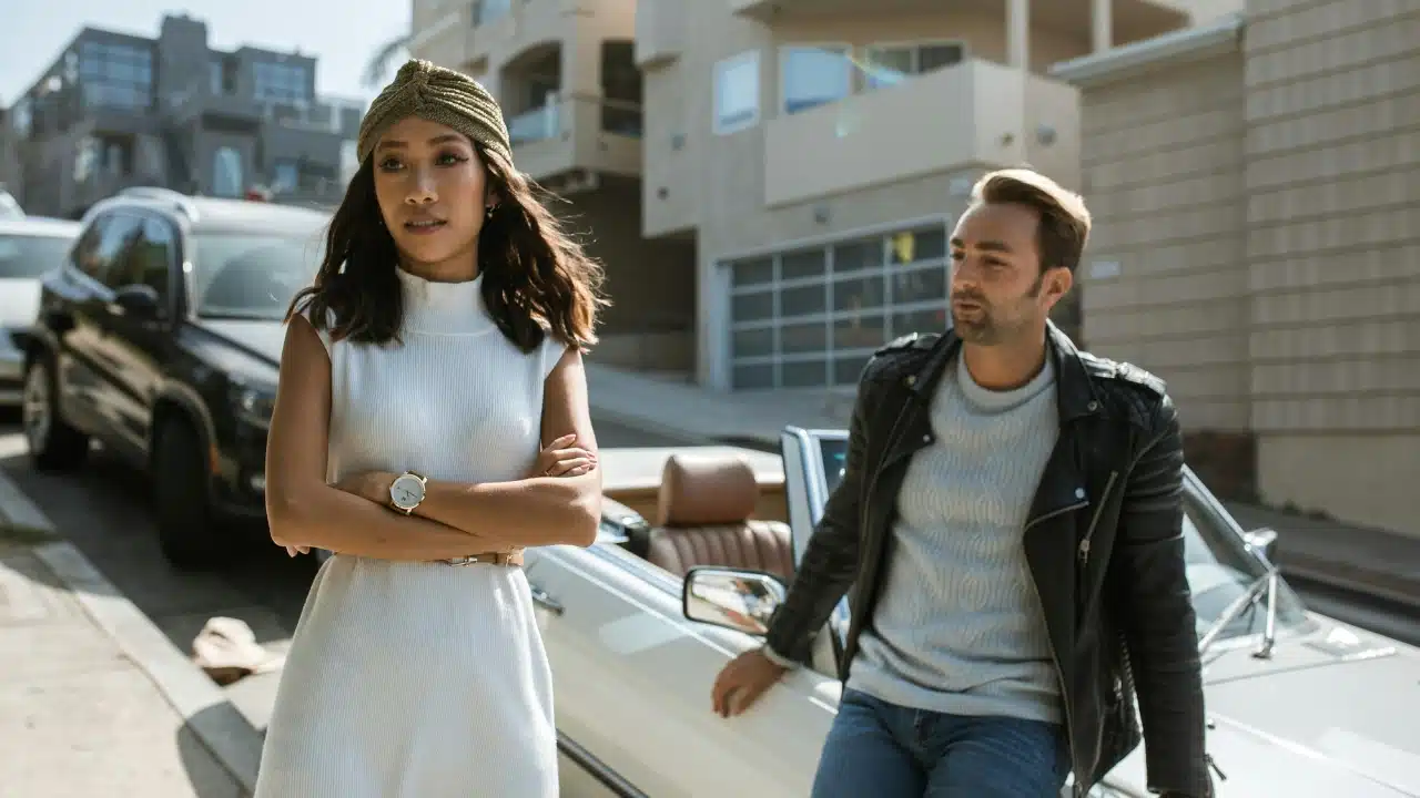 A woman in a white dress stands with crossed arms while a man leans on a car.
