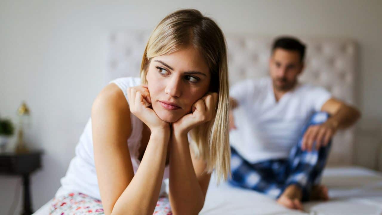 A blonde woman sits on a bed looking away while a man sits behind her.