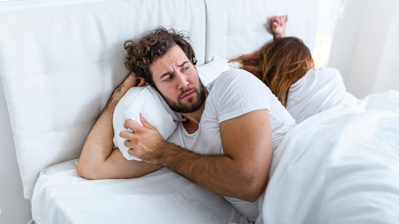 A bearded man lies in bed looking contemplative while a woman sleeps behind him.