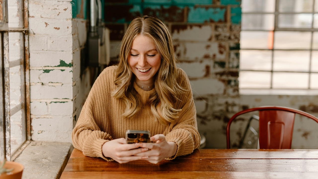 A blonde woman in a brown sweater sits at a table smiling while using a smartphone.