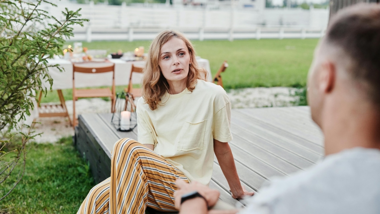 A woman in a yellow shirt and striped pants sits outdoors talking to a man.