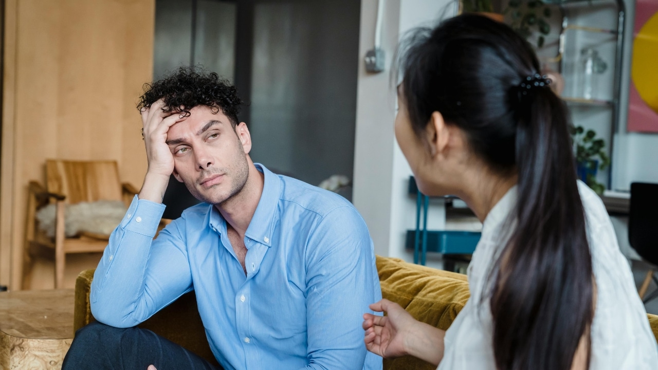 A man sits on a couch with his hand on his head, looking at a woman.